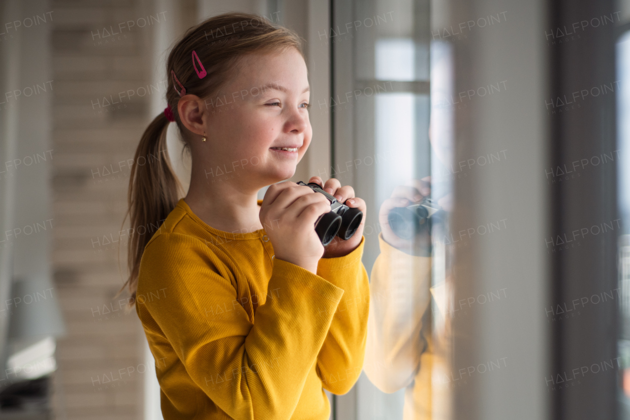 A curious little girl with Down syndrome with binoculars looking through window at home.