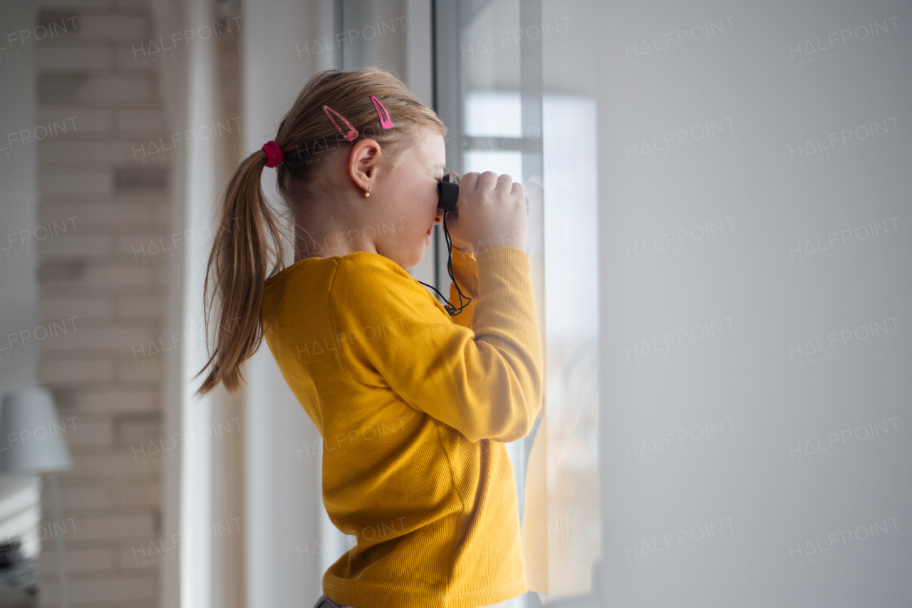 A curious little girl with Down syndrome with binoculars looking through window at home.