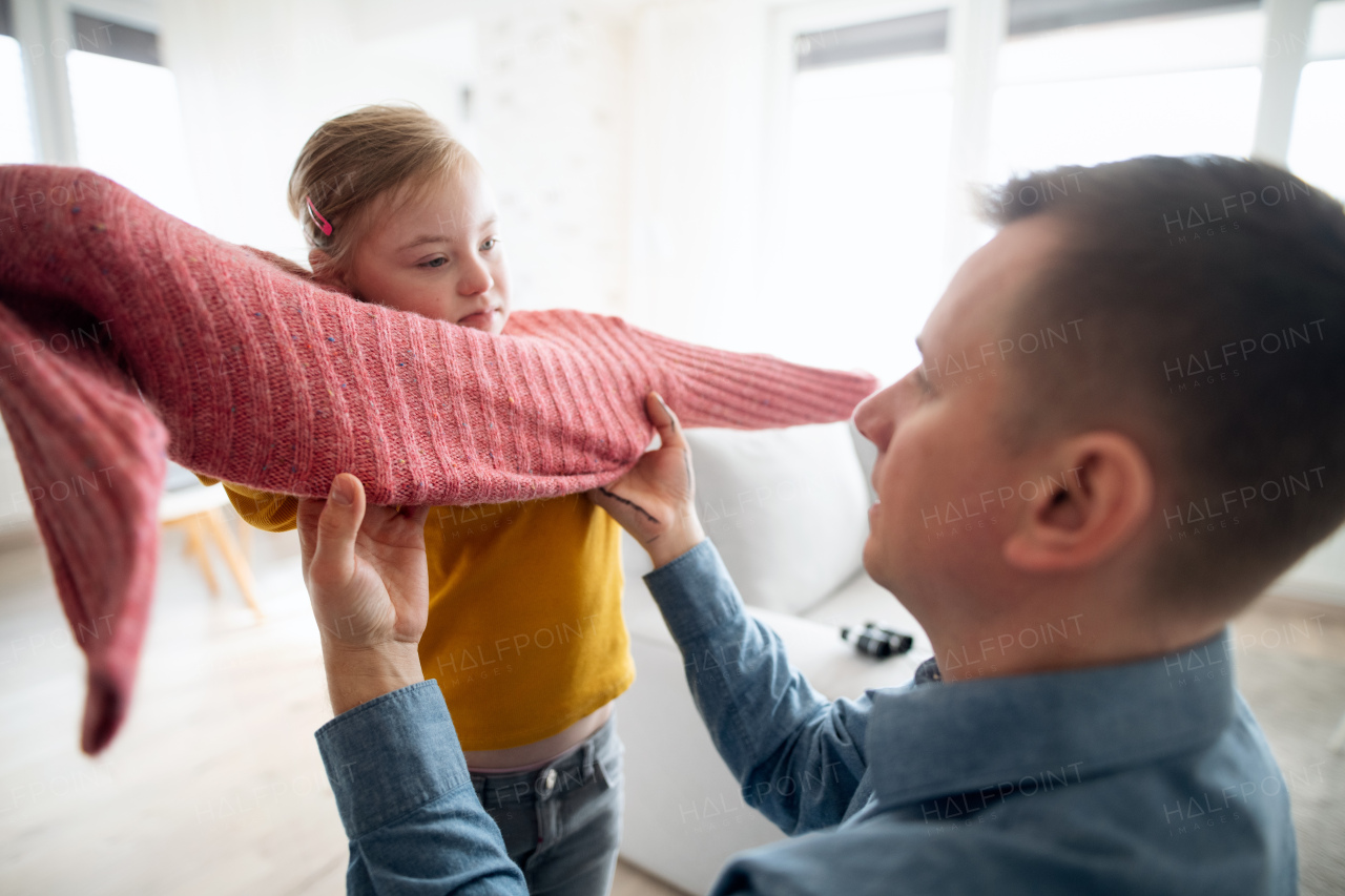 A father helping his little daughter with Down syndrome to wear jumper at home.