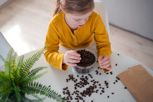 A high angle view of little girl with Down syndrome sitting at table with bowl full of coffee beans at home.