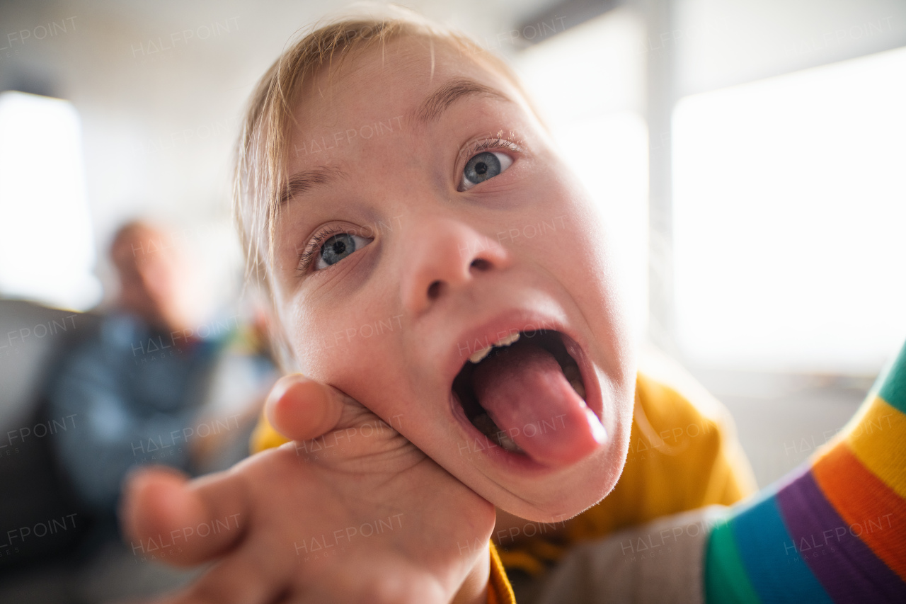 A close-up of cheerful little girl with Down syndrome sticking tongue out at home.