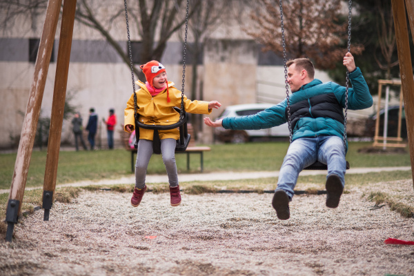 A father with his little daughter with Down syndrome on swings outdoors in playgraound.