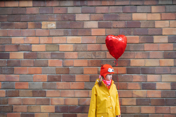 A little girl with Down syndrome holding heart shaped balloon in winter against brick wall.