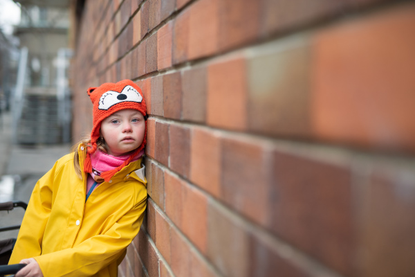 Alittle girl with Down syndrome leaning against brick wall in winter.