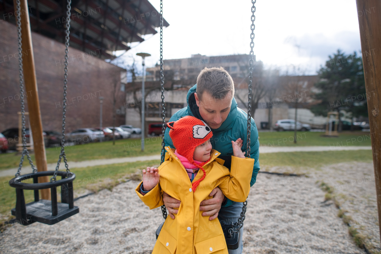 A father pushing his little daughter with Down syndrome on swing outdoors in playgraound.