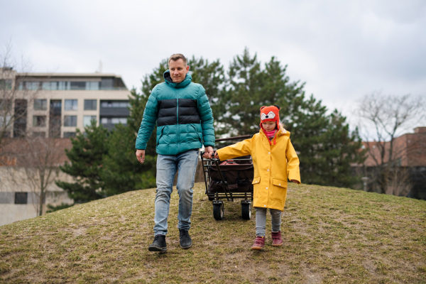 A father with his little daughter with Down syndrome pulling trolley outdoors in park.