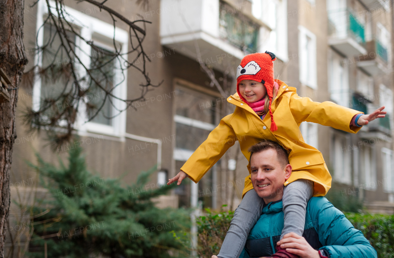 Afather taking his little daughter with Down syndrome on piggyback to school, outdoors in street.