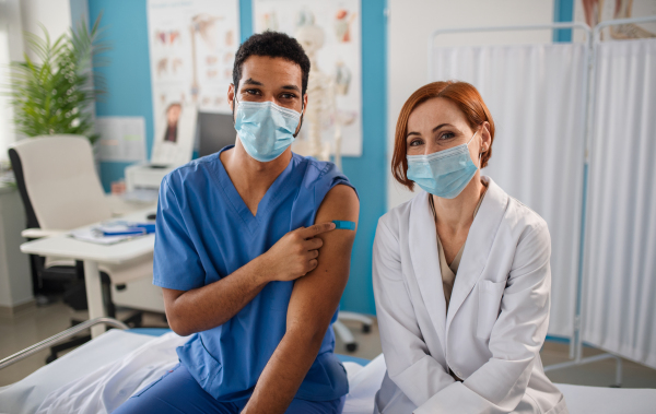 A doctor with colleague wearing medical facemask, looking at camera and showing bandage after vaccine