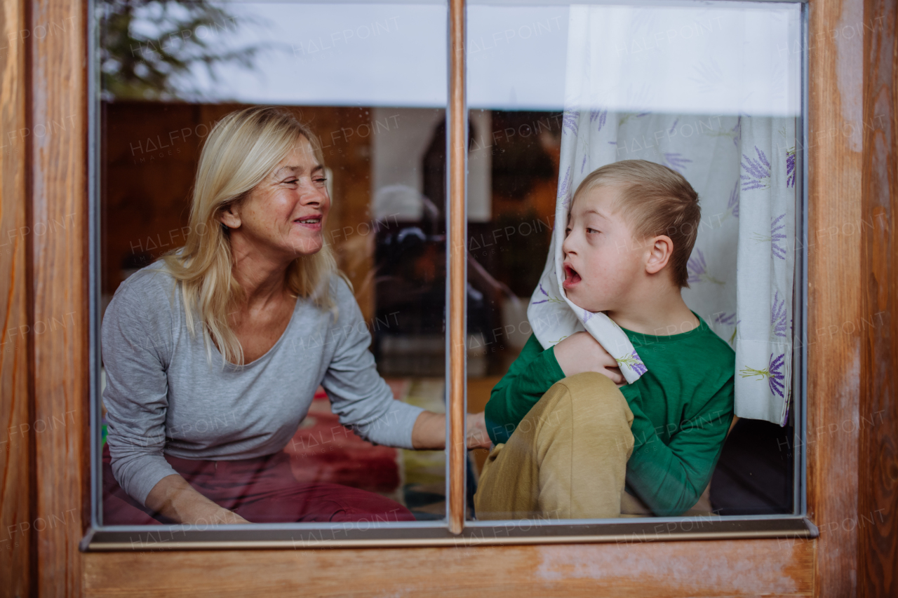 A boy with Down syndrome with his grandmother looking at camera through window at home.