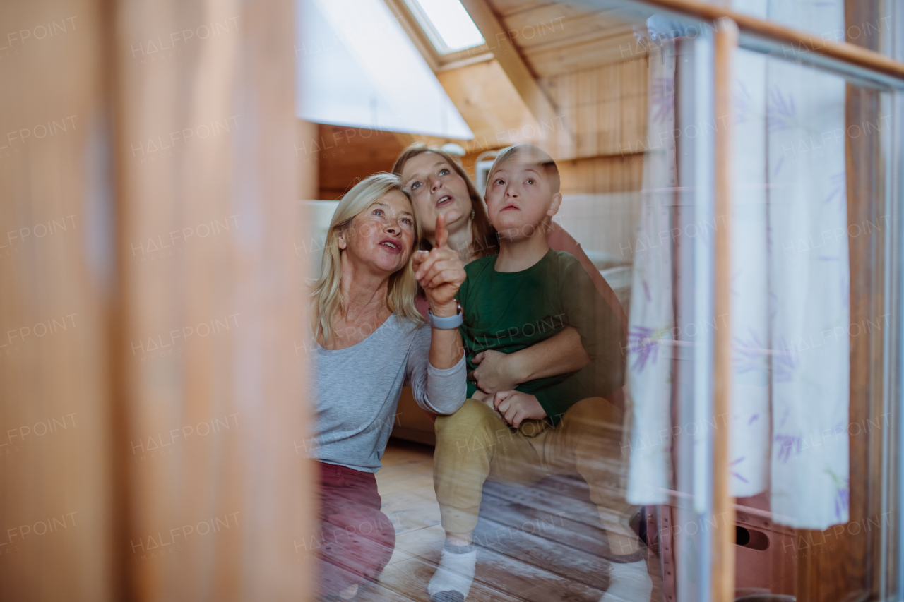 A boy with Down syndrome with his mother and grandmother sitting and looking through window at home.