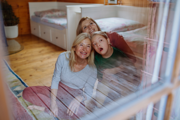 A boy with Down syndrome with his mother and grandmother sitting and looking through window at home.