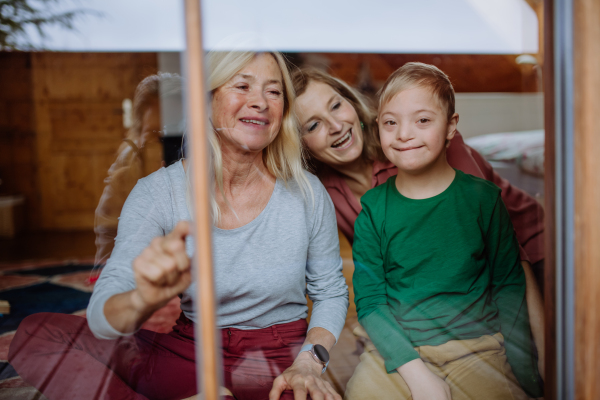 A boy with Down syndrome with his mother and grandmother sitting and looking through window at home.