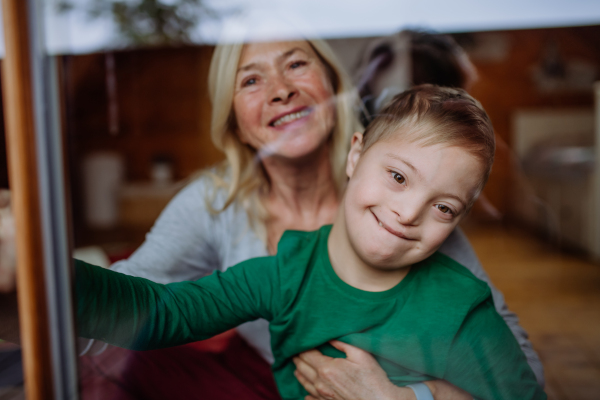 A boy with Down syndrome with his grandmother looking at camera through window at home.