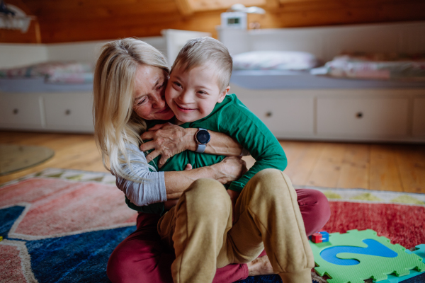 A happy boy with Down syndrome sitting on floor and hugging with his grandmother at home