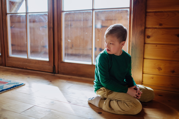 A sad little boy with Down syndrome sitting on floor at home.