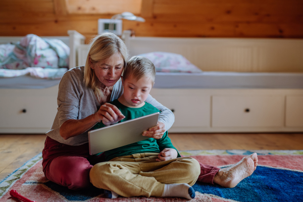 A child with Down syndrome sitting on floor and using tablet with grandmother at home.