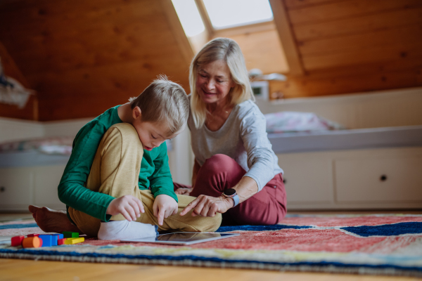 A child with Down syndrome sitting on floor and using tablet with grandmother at home.