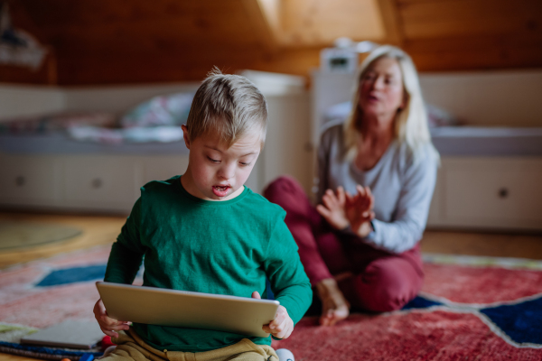 A child with Down syndrome sitting on floor and using tablet with grandmother at home.