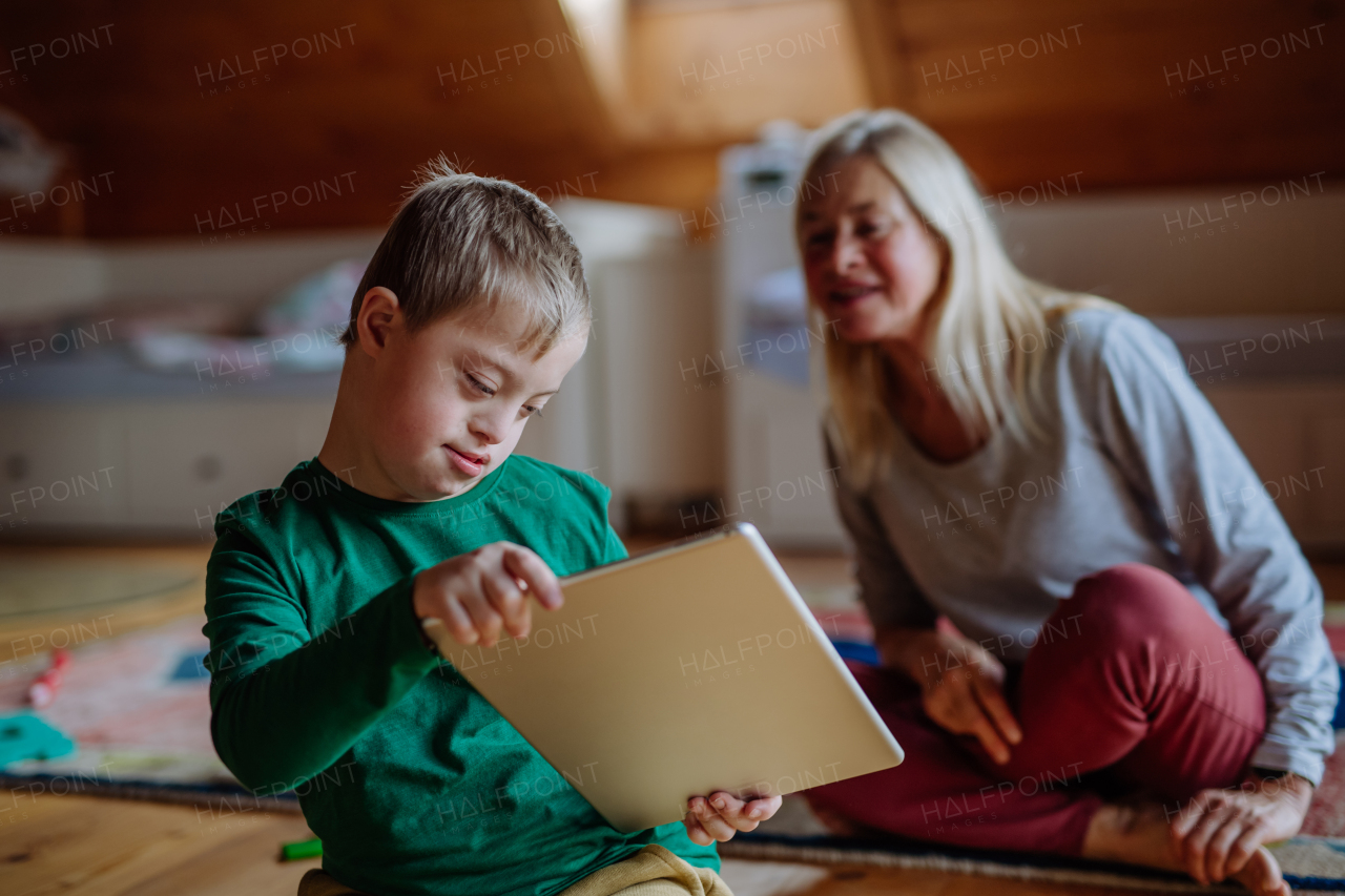 A child with Down syndrome sitting on floor and using tablet with grandmother at home.