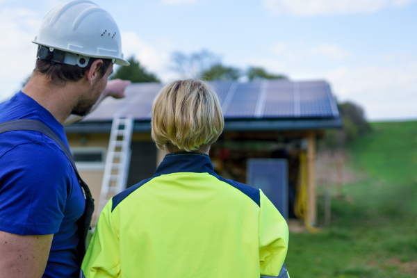 A man and woman solar installers engineers with tablet while installing solar panel system on house, rear view.