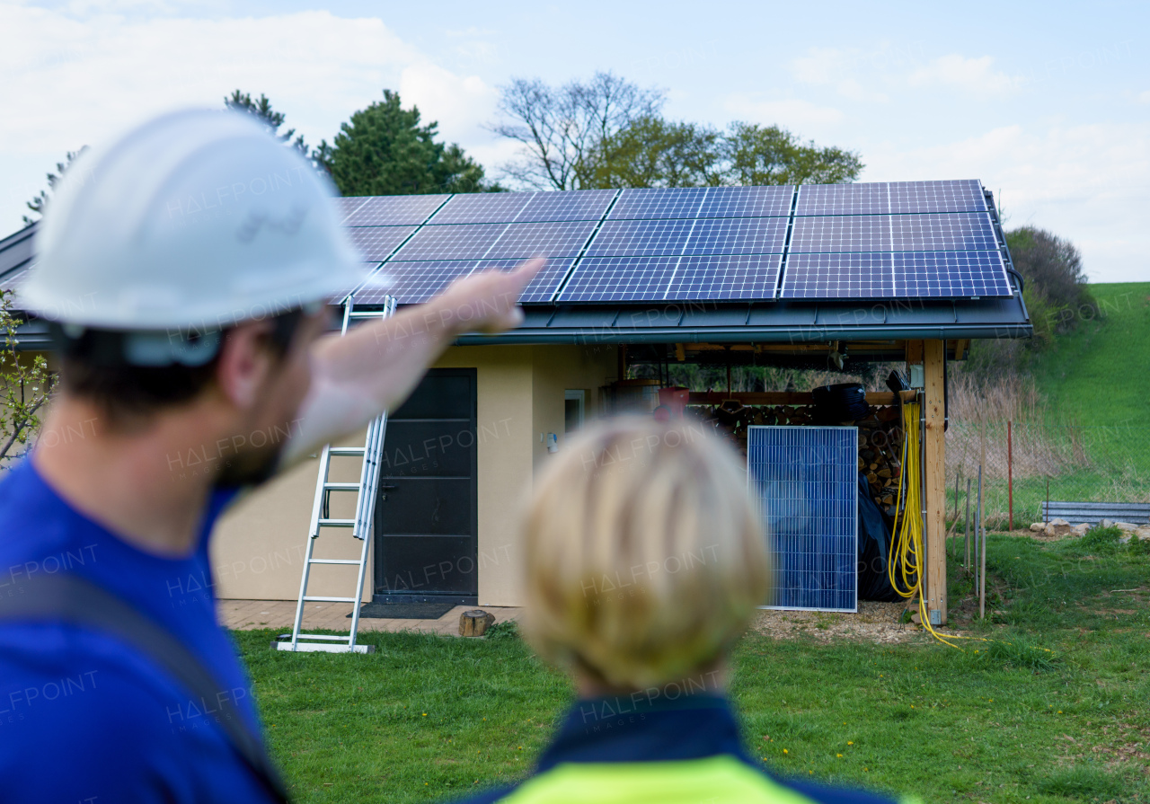 A man and woman solar installers engineers with tablet while installing solar panel system on house, rear view.