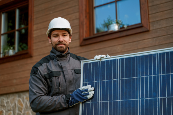 A smiling handyman solar installer carrying solar module while installing solar panel system on house.