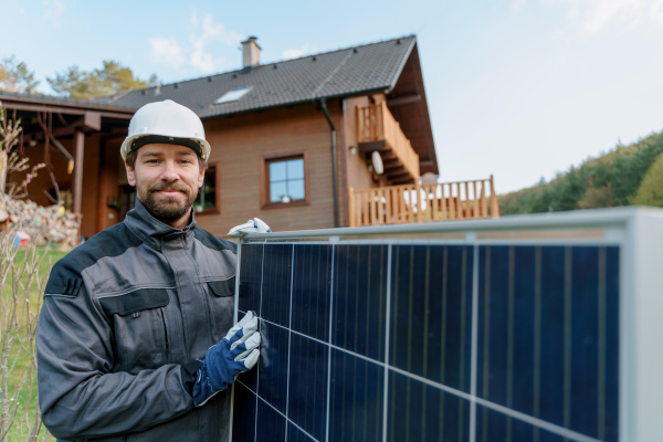 A smiling handyman solar installer carrying solar module while installing solar panel system on house.