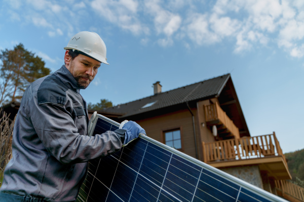 A smiling handyman solar installer carrying solar module while installing solar panel system on house.