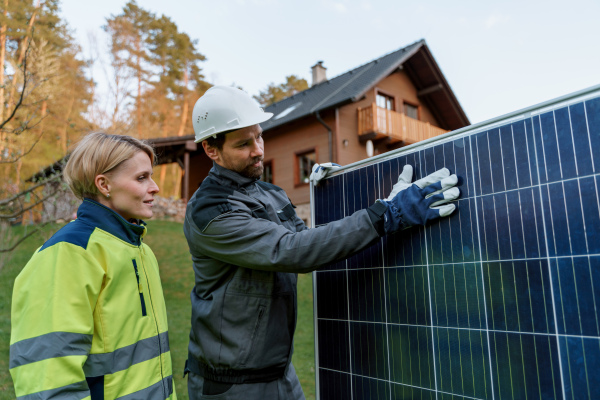 A handyman solar installer discussing with his colleague at solar module in front of family house.