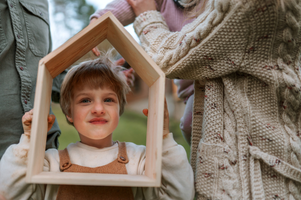 A happy boy with his family is is holidng wooden house frame and looking throug it to camera, new house concept.