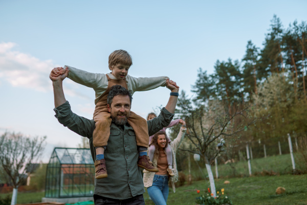 Happy parents with small children on piggybacks running and having fun together in a garden near their house.