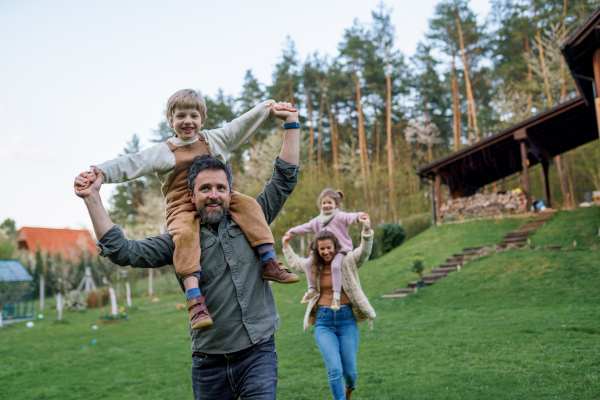 Happy parents with small children on piggybacks running and having fun together in a garden near their house.