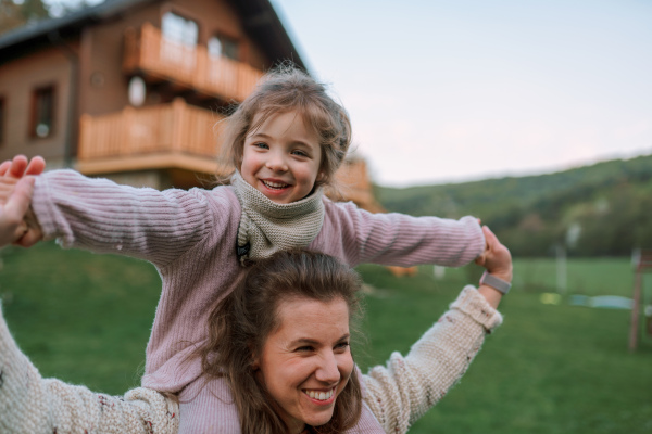 A happy mother with small daughter on piggyback running and having fun together in garden near their house.