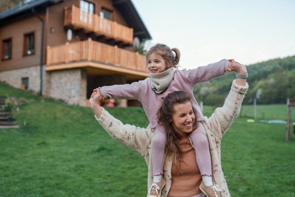 A happy mother with small daughter on piggyback running and having fun together in garden near their house.