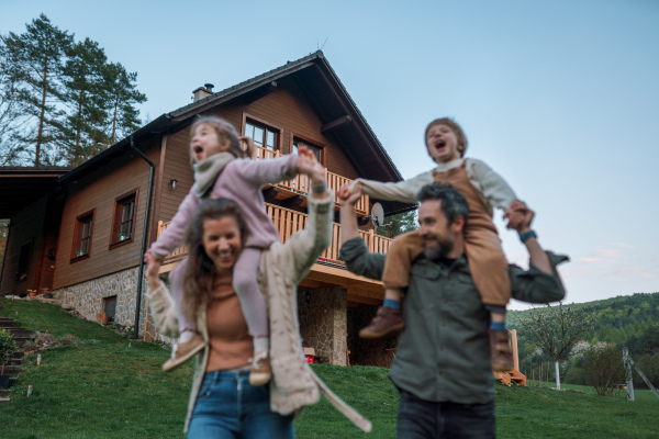 Happy parents with small children on piggybacks running and having fun together in a garden near their house.