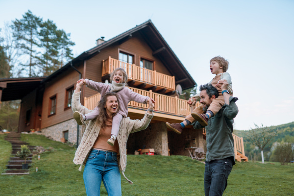 Happy parents with small children on piggybacks running and having fun together in a garden near their house.