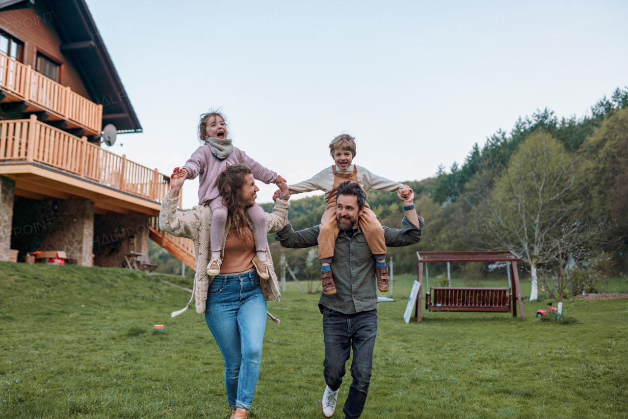 Happy parents with small children on piggybacks running and having fun together in a garden near their house.