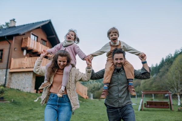 Happy parents with small children on piggybacks running and having fun together in a garden near their house.