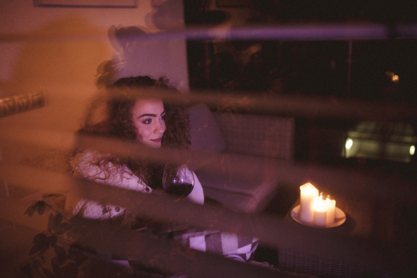 A happy young woman wrapped in blanket sitting on terrace and drinking wine in the evening shot through window.