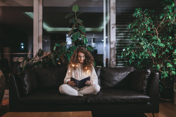 A happy young woman sitting on sofa and reading book in the evening in cozy hyyge living room.