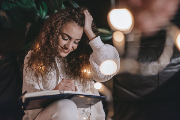 A happy young woman sitting on sofa and writing in diary in the evening in cozy hyyge living room.