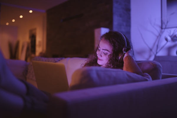 A happy young woman sitting on sofa and litsening to muisc and using laptop the evening in cozy hyyge living room.