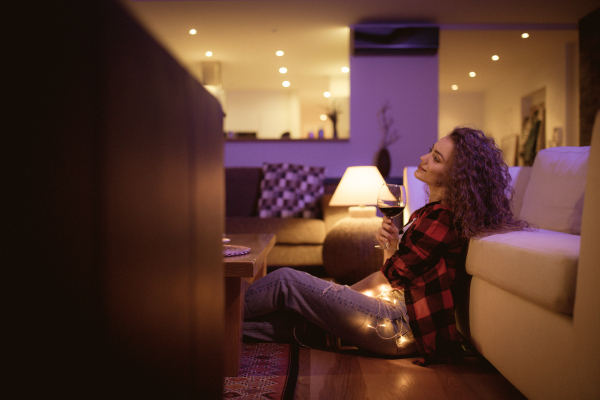 A happy young woman sitting on floor and drinking wine in the evening at home alone.