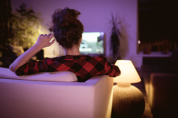 A rear view of single young woman sitting on sofa at home and watching tv.