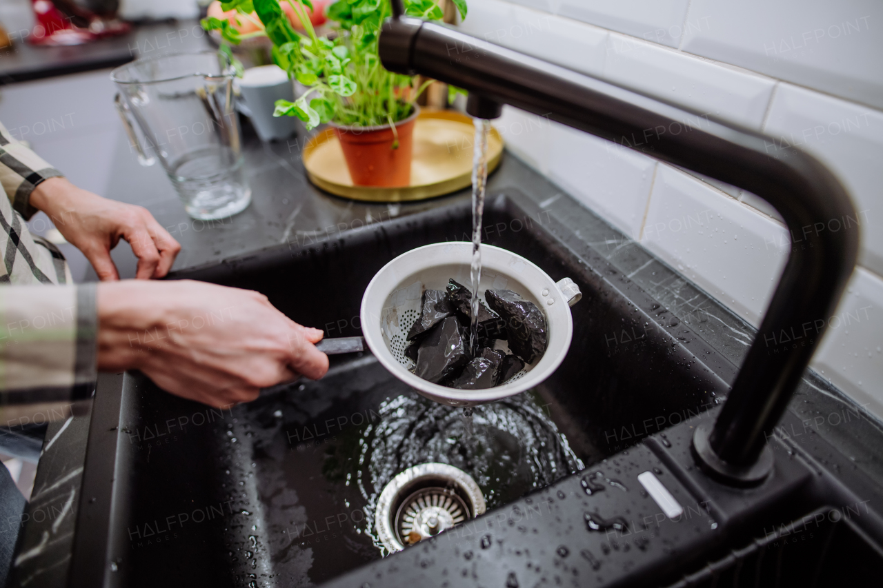 A woman cleaning shungite stones in sieve with pouring water in sink.