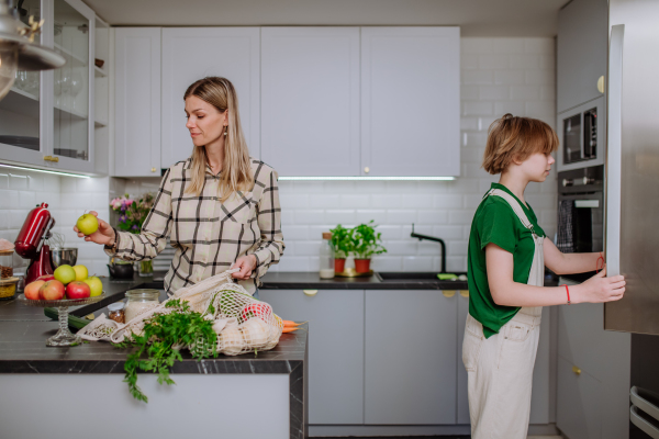 A mother unpacking local food in zero waste packaging from bag with help of daughter in kitchen at home.