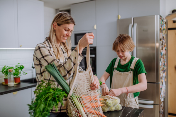 A mother unpacking local food in zero waste packaging from bag with help of daughter in kitchen at home.