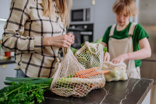 A mother unpacking local food in zero waste packaging from bag with help of daughter in kitchen at home.