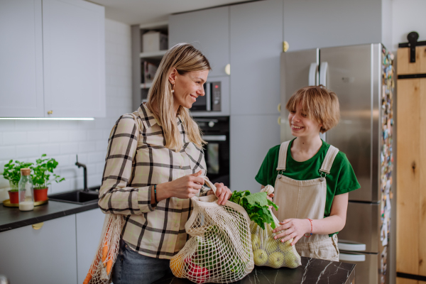 A mother unpacking local food in zero waste packaging from bag with help of daughter in kitchen at home.