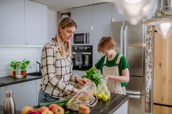 A mother unpacking local food in zero waste packaging from bag with help of daughter in kitchen at home.